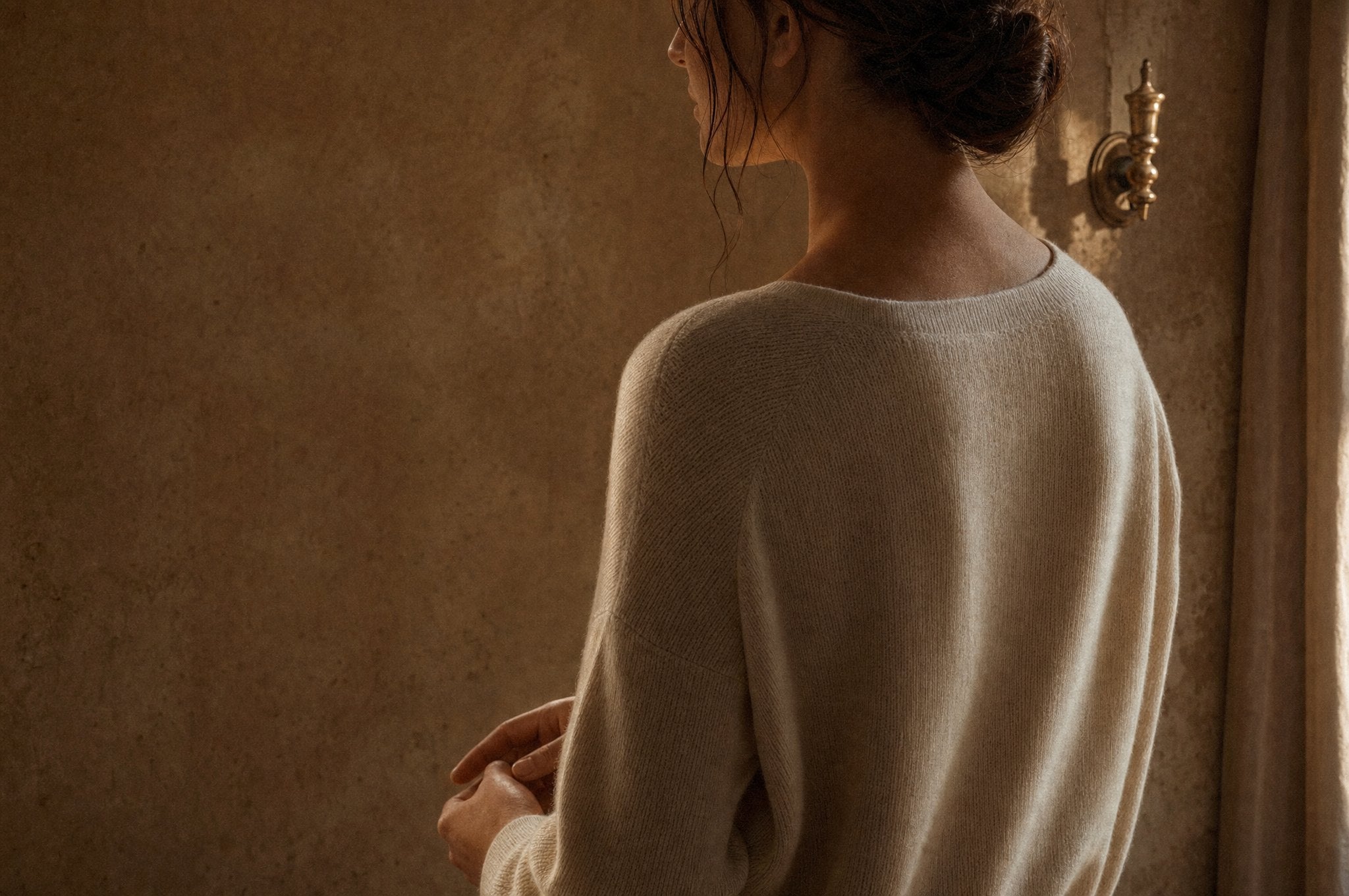 Editorial campaign image: a woman in a cream knit turned towards a warm plaster wall